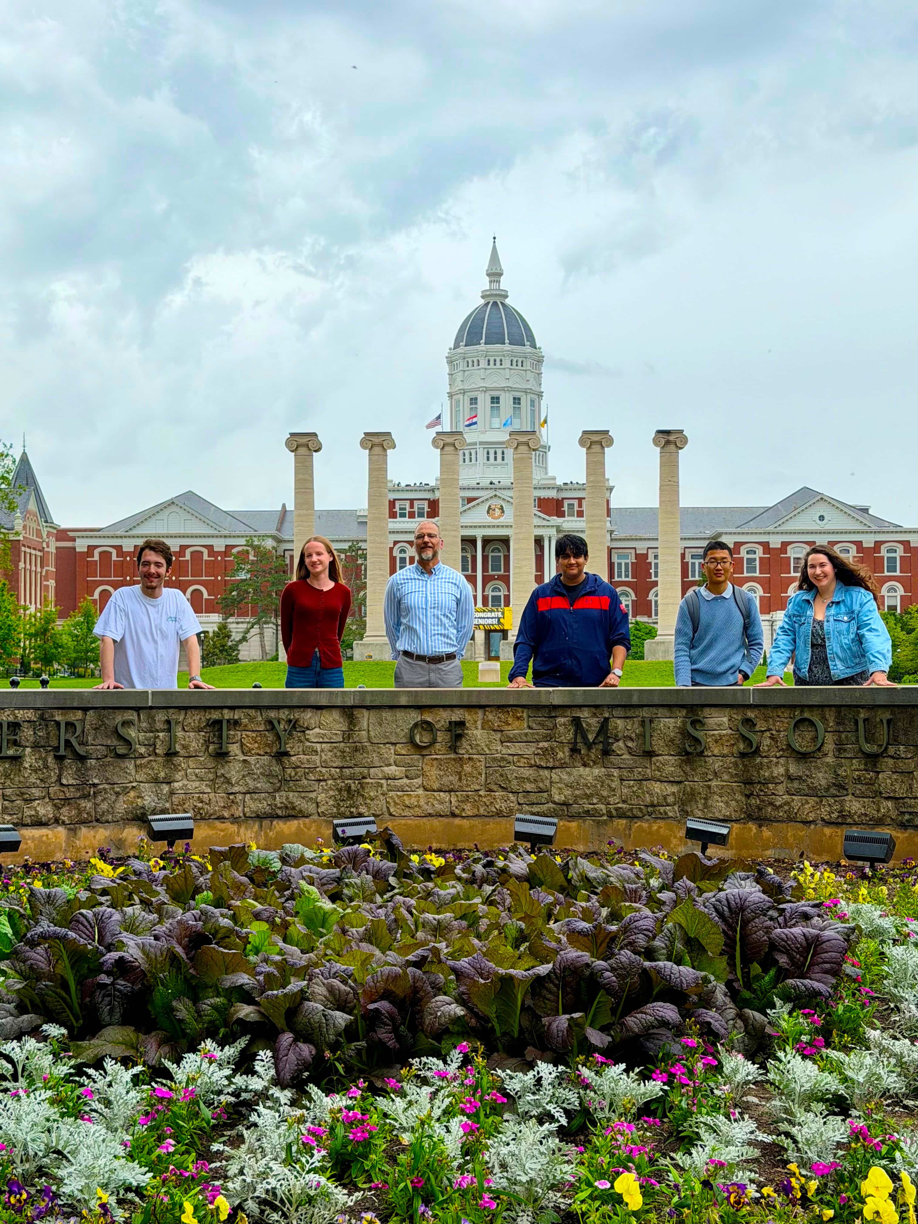 Professor Parsons and graduating students stand smiling behind a "University of Missouri" stone sign, with colorful flowers and the historic campus building with columns in the background on a sunny day.