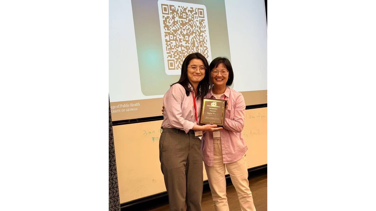 Two people smiling and posing with an award plaque in a classroom setting. A presentation slide with a QR code is displayed in the background.