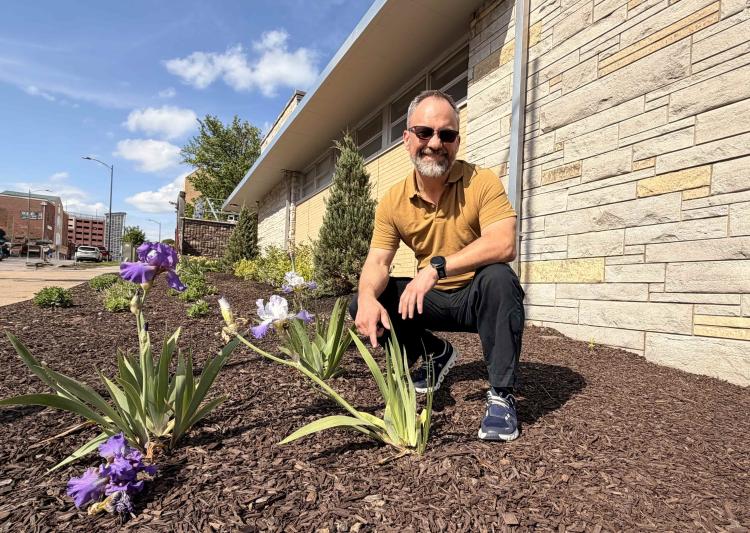 Man in sunglasses and yellow shirt crouches by flowering plants near a modern stone building. Bright day, clear sky, urban backdrop. Smiling.