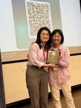 Two people smiling and posing with an award plaque in a classroom setting. A presentation slide with a QR code is displayed in the background.