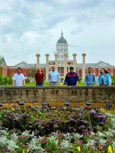 Professor Parsons and graduating students stand smiling behind a "University of Missouri" stone sign, with colorful flowers and the historic campus building with columns in the background.